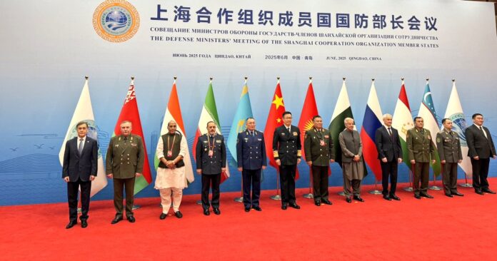 Defense ministers and representatives of Shanghai Cooperation Organisation (SCO) member states stand for a group photograph at the 2025 SCO Defense Ministers' Meeting in Qingdao, China. National flags of the member countries are displayed behind them, including India, China, Russia, Pakistan, Iran, and others. Indian Defence Minister Rajnath Singh is seen in traditional attire near the centre of the lineup. The backdrop displays the SCO emblem and meeting details in Chinese, Russian, and English.