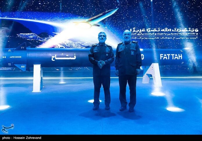 **Alt Text:** Two Iranian military officials stand in front of a displayed Fattah hypersonic missile during its unveiling ceremony, with a digital backdrop showing a missile streaking through space and Persian script above. The scene is illuminated in blue lighting, emphasising the missile and the officials' uniforms.