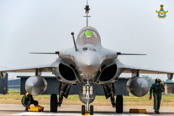 **Alt text:** Front view of an Indian Air Force Rafale fighter jet on the tarmac with two ground crew members performing pre-flight checks; the aircraft's twin air intakes, nose cone, and landing gear are prominently visible. The IAF emblem is seen in the top-right corner.