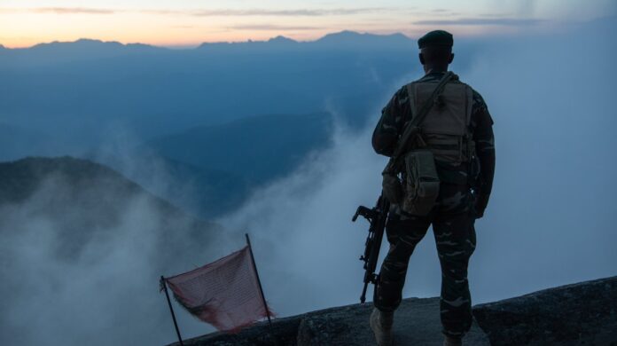 Representative image showing an Indian Army soldier in camouflage gear stands alone on a rocky mountain ridge at dawn, looking out over mist-covered valleys and distant blue peaks. He carries a rifle and a tactical backpack, with a worn red prayer flag fluttering nearby. The scene evokes silence, vigilance, and the harsh terrain typical of high-altitude warfare, reminiscent of the Kargil conflict.